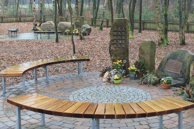 Abgebildet ist der Wald in Altenwahlingen. Es ist eine Gedenkstätte mit Bänken ist zu sehen. Blumen stehen vor Gedenksteinen.